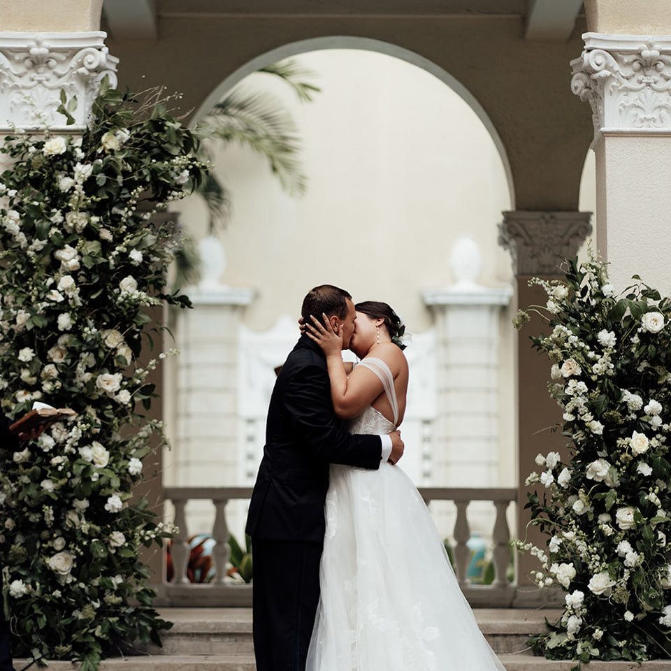 Bride and Groom kissing under ceremony arch of white and green florals at Cafe Julia, Hawaii designed by SF.Fleur