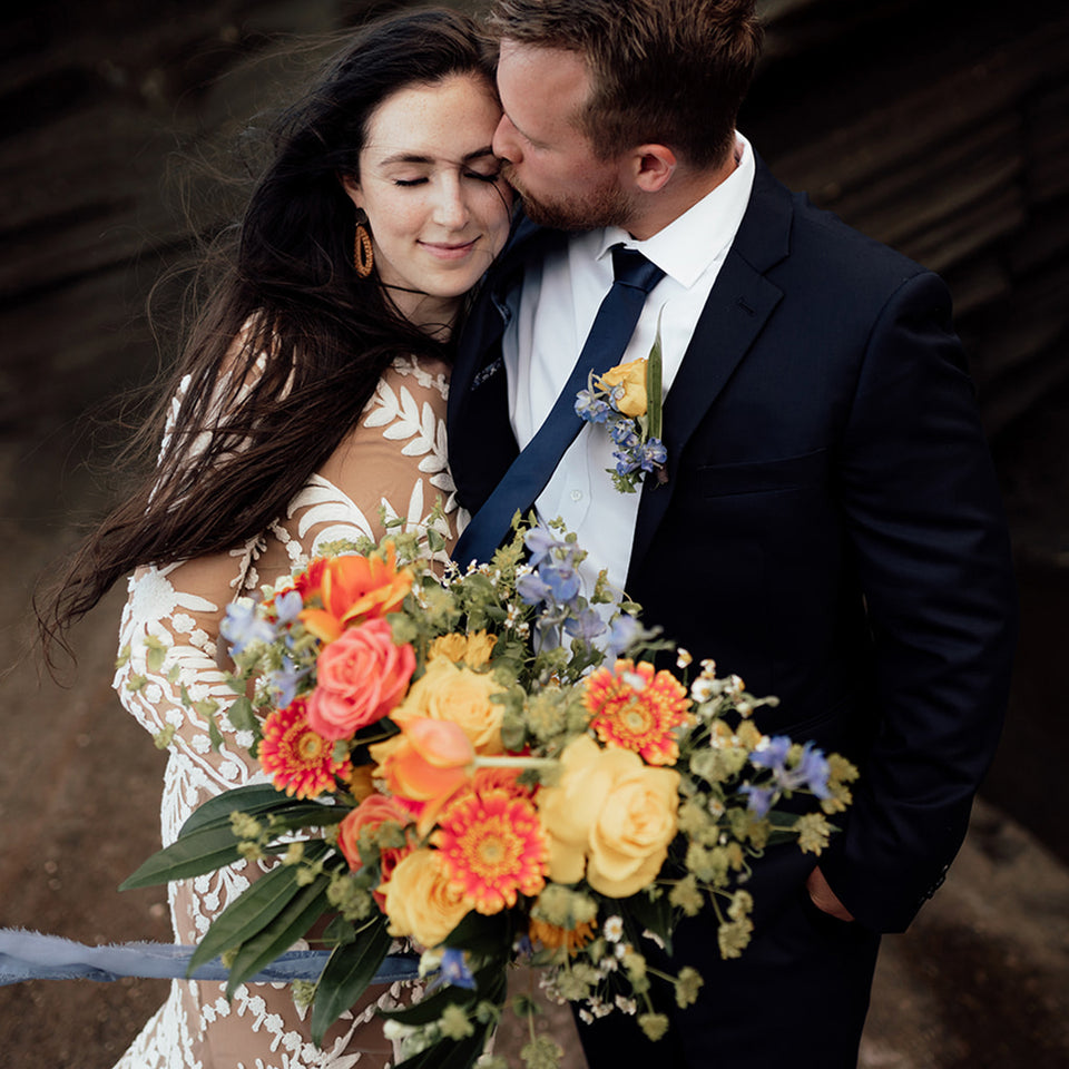 Bride being hugged by groom and kissed on foreheard while holding a colorful orange and yellow bouquet designed by SF.Fleur.